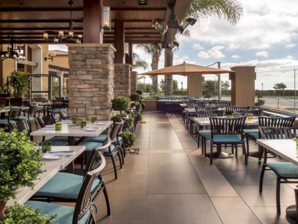 A stylish outdoor restaurant or cafe with stone pillars, turquoise cushioned chairs, white tables, potted plants, and shaded seating facing a waterfront view.