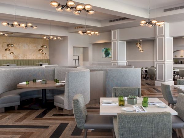 A modern, airy restaurant with gray upholstered booths, round and square tables, soft lighting, and geometric wood flooring, creating a chic dining space.