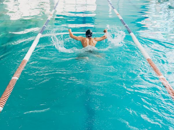 A swimmer is mid-dive in a turquoise pool, arms spread, streamlined between lane ropes, making a powerful splash as they glide forward.