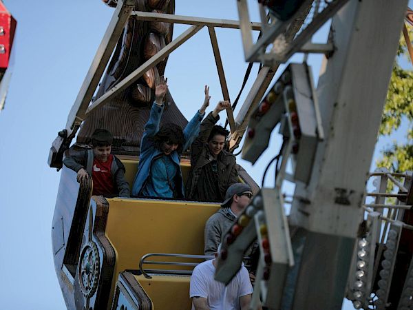 Three people ride a ferris wheel car high above the ground, raising their hands as the ride looms against a clear sky.