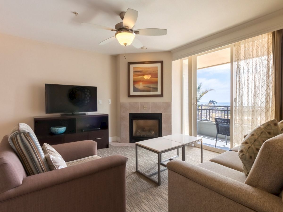 Cozy living room with two beige sofas, a TV on a stand, a fireplace, framed art, and a sliding glass door opening to a balcony with ocean view, bright natural light.